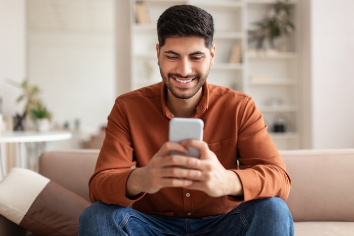 A person in jeans and a button-down shirt sits on a couch smiling at a phone in his hands.